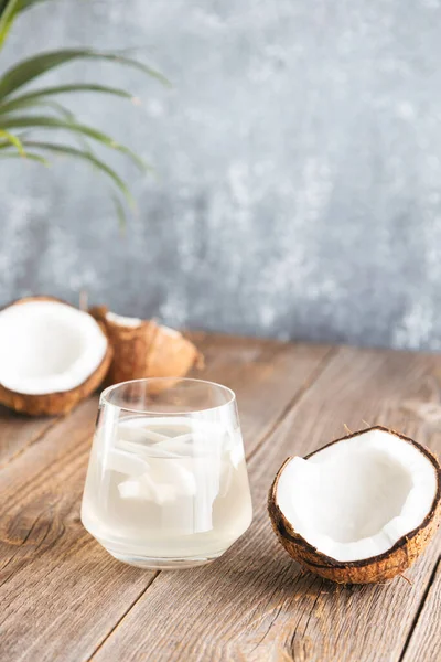 Ein Glas frisches, biologisches Kokoswasser, Milch auf einem Holztisch unter Palmblättern. Gehackte saftige Kokosnüsse liegen in der Nähe. Vertikales Foto — Stockfoto