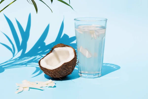 Água de coco em um copo com um tubo de coquetel e meio coco fresco perto em um fundo azul abaixo de folhas de palma em um dia ensolarado. Sumo de desintoxicação perfeito — Fotografia de Stock