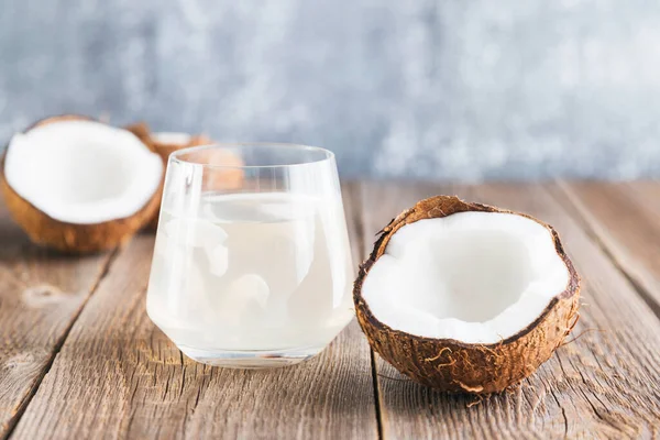 W pobliżu znajduje się woda kokosowa w szklanym zbliżeniu na drewnianym stole i pół świeżego orzecha kokosowego. Organiczny sok kokosowy — Zdjęcie stockowe