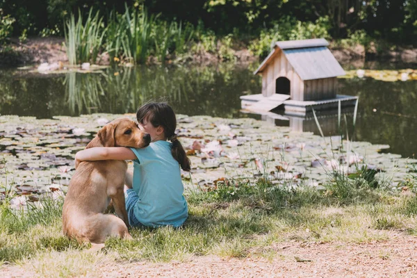 Friendship of animals and children, people. Caucasian girl sits on the bank of a pond, river and hugs a labrador puppy. Outdoor games with animals — Stock Photo, Image