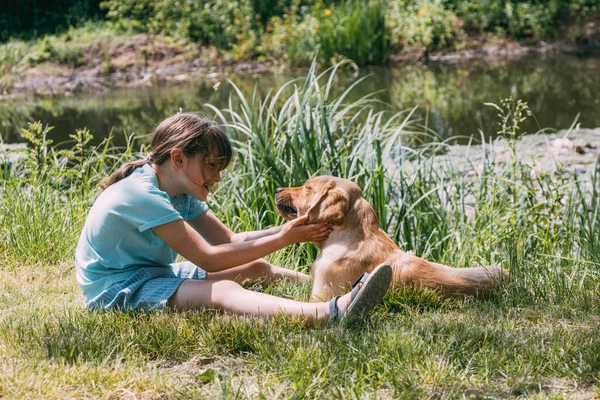 Přátelství zvířat a dětí, lidí. Běloška sedí na břehu rybníka, řeka a hraje si se psím štěnětem, labradorem retrívrem. Venkovní hry, letní dovolená — Stock fotografie