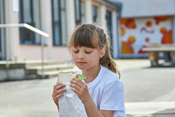 Portrait d'une écolière qui mange son déjeuner, une collation dans la cour de l'école. Nourriture pour les enfants dans les établissements d'enseignement, jardins d'enfants. — Photo