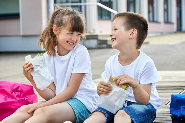 Schoolchildren boy and girl laugh and eat their lunch, snack, breakfast in the school yard. Food for children in educational institutions, kindergartens. — Stock Photo, Image