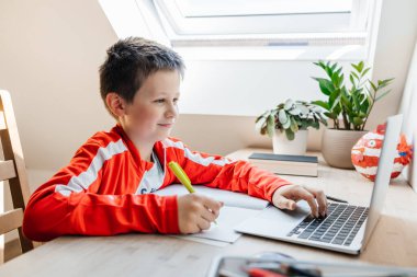 Genç bir çocuk açık bir dizüstü bilgisayarla ders çalışıyor ve masasına notlar yazıyor.