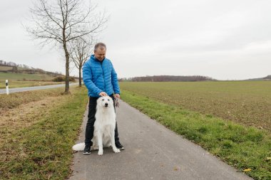 Bir adam kışın kırsal bir yolda köpeğini okşayarak dışarıda vakit geçirir.
