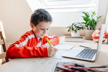 Genç bir çocuk açık bir dizüstü bilgisayarla ders çalışıyor ve masasına notlar yazıyor.