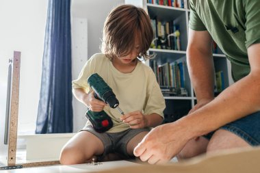 Küçük bir çocuk babasından diy becerisi öğreniyor, matkapla mobilya yapıyor ve vida yapıyor.