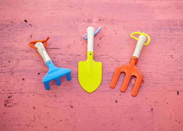 A set of children\'s small gardening tools including a spade, digger and rake on a pink wooden background