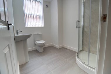 Yeni monte edilmiş tuvalet ve lavabo, fayanslarla döşenmiş gri zemin, beyaz duvarlar ve penceresinde panjurlar olan yeni bir İngiliz tuvaletinin fotoğrafı.