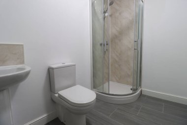 Yeni monte edilmiş tuvalet, lavabo ve duş kabini, fayanslı gri zemin, beyaz duvarlar ve etek tahtaları olan modern bir İngiliz tuvaletinin fotoğrafı.