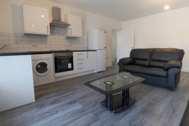 Modern bir İngiliz apartmanı oturma odası ve mutfağı. Siyah deri koltuğu ve gri ahşap zemini olan bir sehpası var. Beyaz duvarları var..