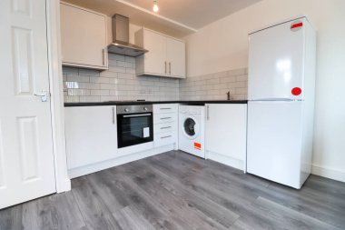 Beyaz duvarları olan, gri ahşap zeminli modern mutfak çamaşır makinesi, fırın ve beyaz buzdolabıyla donatılmış modern bir İngiliz daire mutfağı..