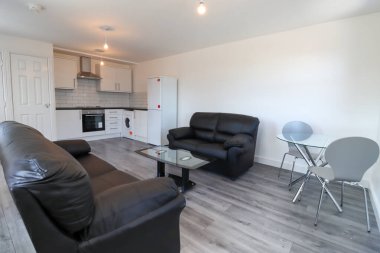 Modern bir İngiliz apartmanı oturma odası ve mutfağı. Siyah deri koltuğu ve gri ahşap zemini olan bir sehpası var. Beyaz duvarları var..