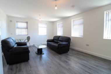 Modern bir İngiliz oturma odası, iki siyah deri koltuğu ve gri ahşap zeminde bir yemek masası, beyaz duvarları ve penceresinde modern panjurları olan.