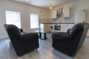 Modern bir İngiliz apartmanı oturma odası ve mutfağı. Siyah deri koltuğu ve gri ahşap zemini olan bir sehpası var. Beyaz duvarları var..