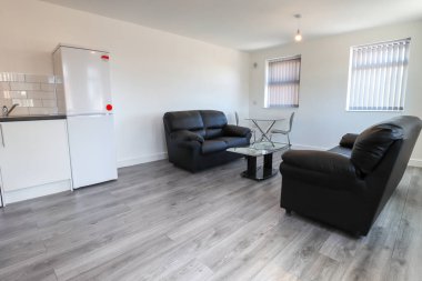 Modern bir İngiliz apartmanı oturma odası ve mutfağı. Siyah deri koltuğu ve gri ahşap zemini olan bir sehpası var. Beyaz duvarları var.