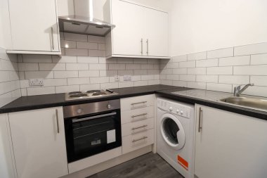 Gri ahşap zemini, beyaz duvarları, çamaşır makinesi, fırını ve mutfak lavabosu olan modern bir İngiliz mutfağı.