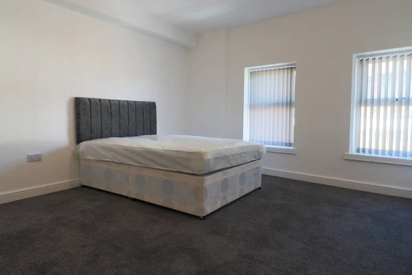 A newly decorated typical British UK bedroom with a large bed and wardrobe, with white walls, grey carpet with blinds at the window.