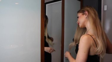 Siyah elbiseli, uzun sarı saçlı, çekici bir kadın. Saçını aynada tarıyor, hazırlanıyor ya da bir randevuya çıkıyor.