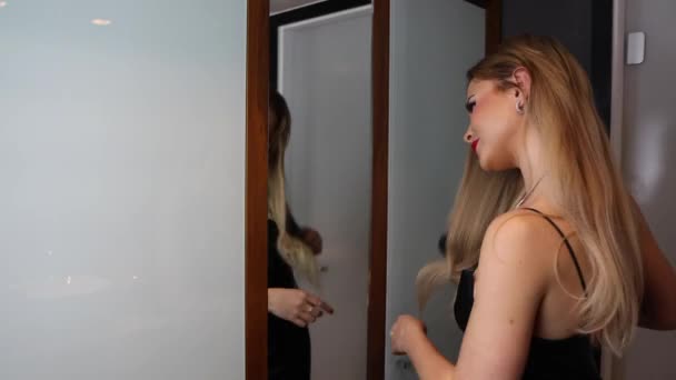 一个漂亮的女人 一头长长的金发 穿着一件黑色的衣服 看起来很优雅 她在镜子里用梳子梳理她的头发 为约会做准备 — 图库视频影像