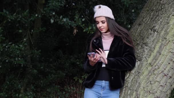 公園で若い魅力的な女性は冬の時間に木にもたれて 彼女の電話をオンラインで見て日付左と右にスワイプ日付アプリ プロファイルのいくつかを好み いくつかを好きではありません — ストック動画