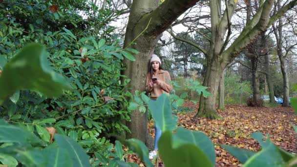 겨울에 핸드폰을 사용하는 여성은 공원에서 점퍼와 모자를 누군가가 나뭇가지와 덤불을 — 비디오