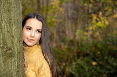 Kışın uzun kahverengi saçlı çekici bir kadın ahşap bir parkta ağaçların arkasından dışarı bakan sarı bir kazak giyiyor..