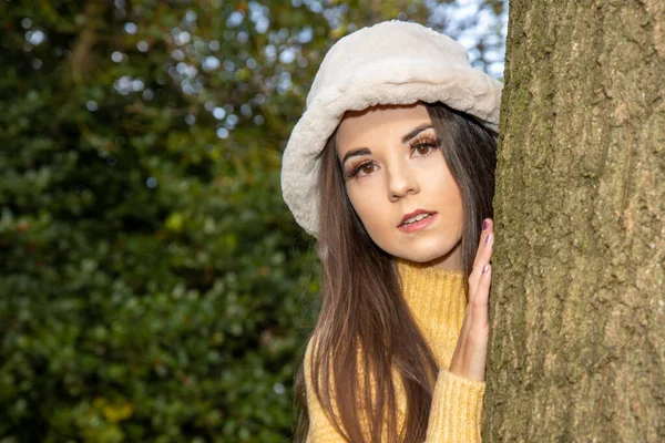 A young attractive woman with long brown hair in the winter time in a wooden park area wearing a yellow jumper looking out from behind a tree.