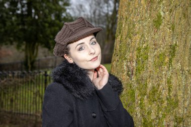 A attractive young woman in a public park on a cold day in the winter time looking sexy and seductive while leaning up against a tree