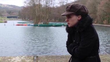 Parkta kışın göl kenarında kalın siyah ceketli ve örgü şapkalı birini beklerken soğuk ve üzgün görünen çekici bir genç kadın.
