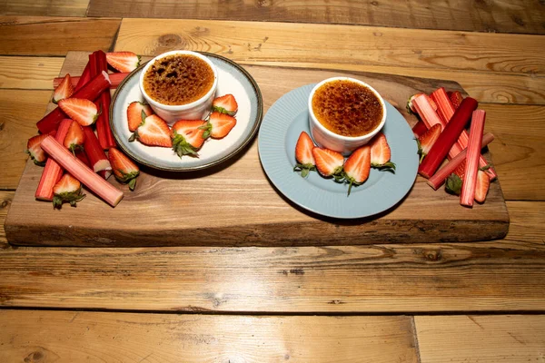 A delicious french desert of Rhubarb and Cardamom Creme Brulee on a wooden kitchen table surrounded by strawberries and Rhubarb