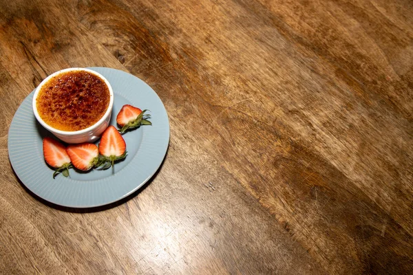 A delicious french desert of Rhubarb and Cardamom Creme Brulee on a wooden kitchen table with cut strawberries on the side