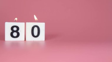 80 numaranın bir doğum günü veya yıldönümü kutlaması için pembe arka planda yanıp söndüğünü söyleyen kare bir mum.