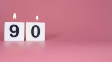 90 numaranın bir doğum gününü veya yıldönümünü kutlayan pembe bir arka planda yanıp söndüğünü söyleyen kare bir mum.