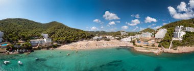 İspanya 'nın İbiza adasının geniş açılı panoramik fotoğrafı yazın güneşli bir günde Cala Llonga sahilindeki güzel plaj ve tatil otellerini gösteriyor.
