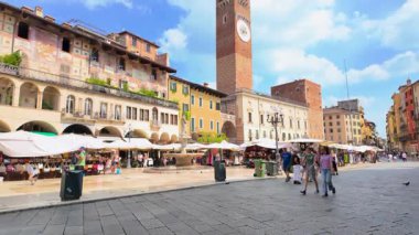 Verona, İtalya - 30 Ağustos 2025: Güneşli bir yaz gününde büyük bir saat kulesinin yanından geçen insanları ve turistleri gösteren güzel Verona şehrinin yavaş çekim görüntüleri