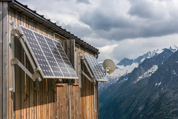 PV solar panels and satellite dish antenna at the wall of a wooden building with snowy mountains in the background.