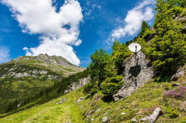 Uydu anteni yeşil dağların etrafındaki bir kayanın üzerine monte edilmiş. Güzel yaz dağ manzarası - beyaz bulutlu mavi gökyüzü.