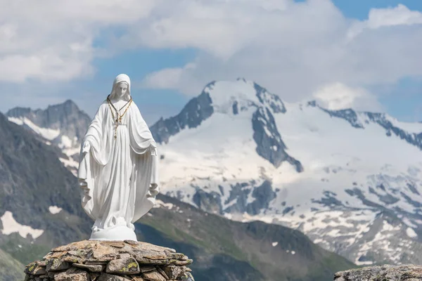 White statue of Virgin Mary, Mother of God, placed on top of the mountain. In the background there are snowy peaks of high mountains, blue sky, white clouds.