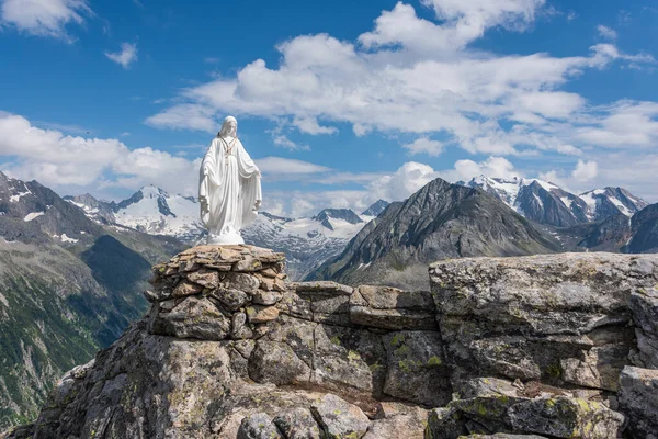 White statue of Virgin Mary, Mother of God, placed on top of the mountain. In the background there are snowy peaks of high mountains, blue sky, white clouds.