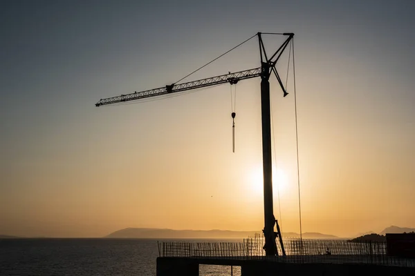 Silhouette, outline of the crane on the construction site against the setting sun and the sea. A fragment of the building, ribbed walls and reinforcement visible in the foreground