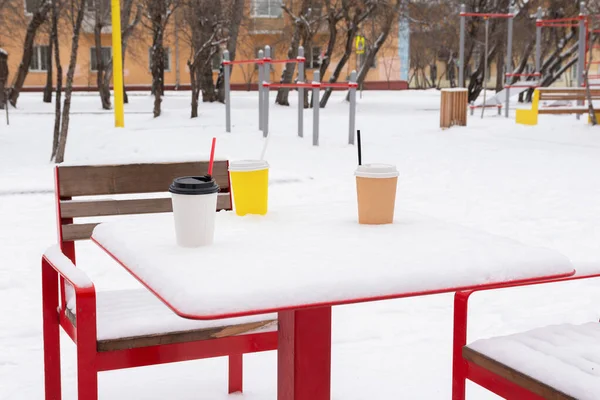 Three cups of coffee are on a snow-covered table in a winter park.