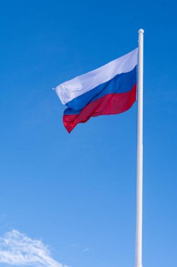 Rusya bayrağı, mavi gökyüzüne karşı uzun bir bayrak direğinde rüzgarda sallanıyor..