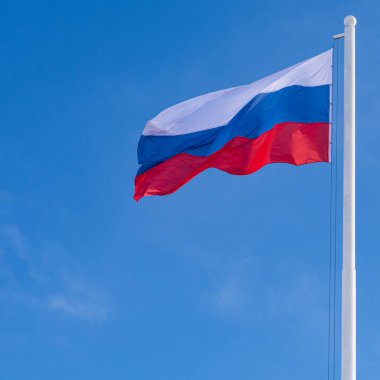 Rusya bayrağı, mavi gökyüzüne karşı uzun bir bayrak direğinde rüzgarda sallanıyor..