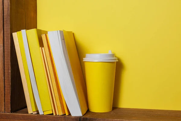 Books with yellow covers and a paper coffee cup sit on a brown old wooden shelf.