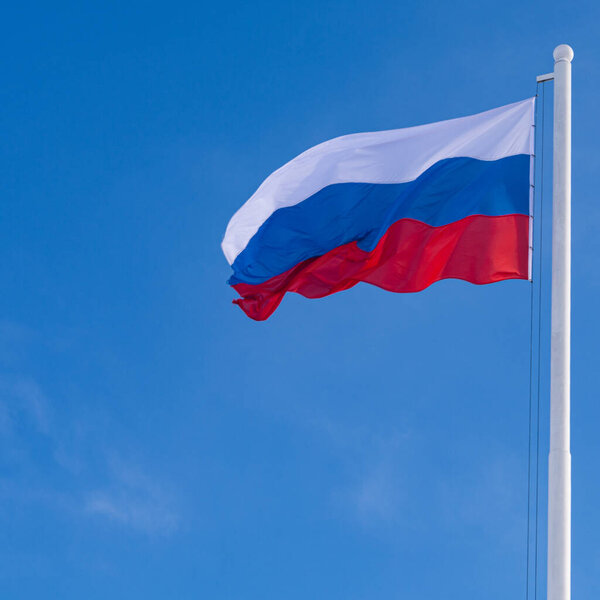 The flag of Russia waving in the wind on a long flagpole against the blue sky.
