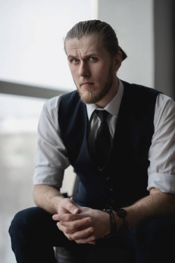 Beyaz gömlekli ve yelekli bir adam koltuğa oturur ve kameraya poz verir.