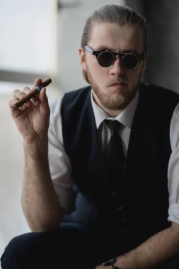 Beyaz gömlekli ve yelekli bir adam elinde sigarayla kanepede oturuyor.