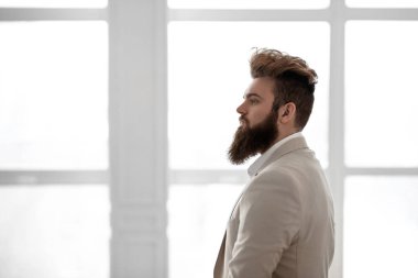 Pencerenin önünde beyaz takım elbiseli sakallı bir adam.