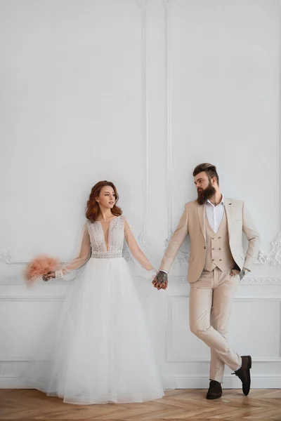 Beyaz elbiseli bir adam ve bir kız beyaz bir duvara yaslanıp el ele tutuşuyorlar.